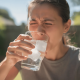 A Person Is Wincing In Discomfort While Sipping A Glass Of Ice Water On A Sunny Day, Indicating Potential Tooth Sensitivity Or Pain.