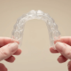 Invisalign: A Clear Choice For A Healthier, More Confident Smile 12 A Clear Aligner Tray Is Being Held Between Two Hands Against A Neutral Background, Showcasing Its Discreet Design As A Modern Orthodontic Treatment Option For Achieving Straighter Teeth.